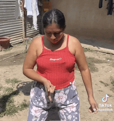 a woman in a red tank top and floral pants is standing in a dirt yard .