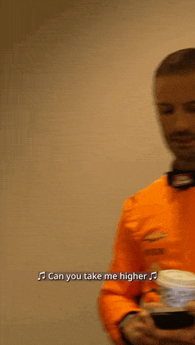 a man in an orange shirt is holding a cup of coffee and singing " can you take me higher "