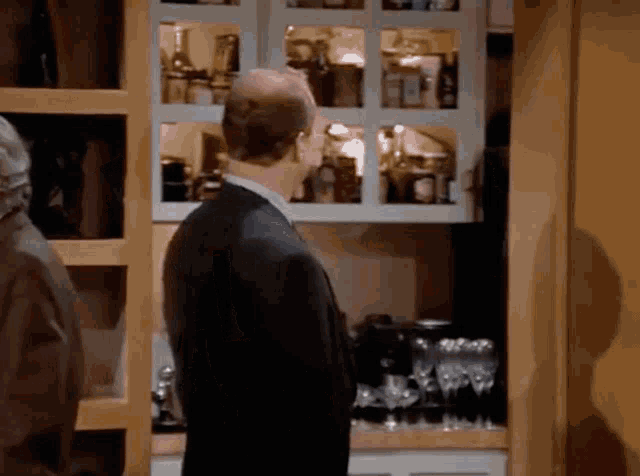 a bald man in a suit stands in front of a shelf full of bottles
