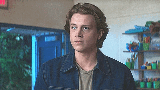 a young man in a denim jacket stands in a room with a red exit sign behind him