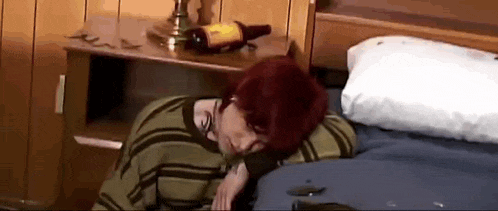 a person with red hair is laying on a bed with a bottle of wine on the nightstand .