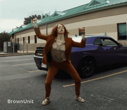 a woman in a suit is dancing in front of a purple dodge challenger car