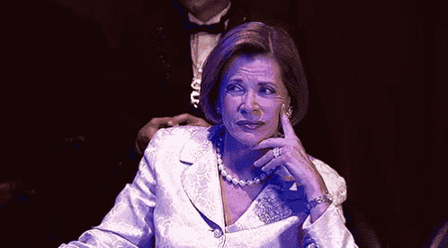 a woman in a white suit and pearl necklace is sitting in a dark room with a man in a tuxedo .