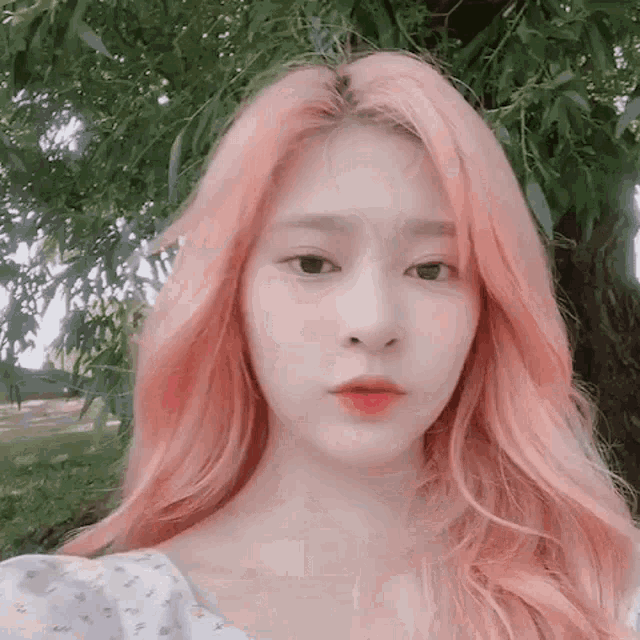 a girl with pink hair is standing in front of a tree and looking at the camera .