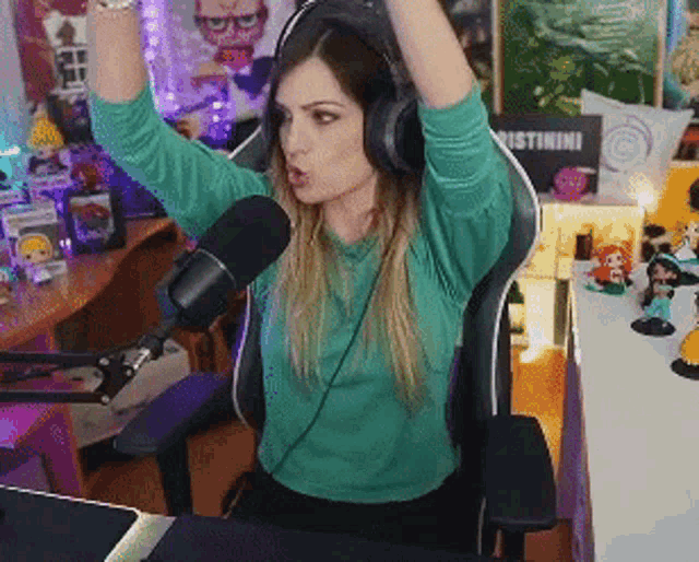 a woman wearing headphones and a green shirt is sitting in front of a microphone with her hands in the air .
