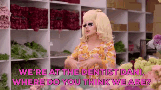 a drag queen is standing in front of a shelf full of flowers and talking to a dentist .