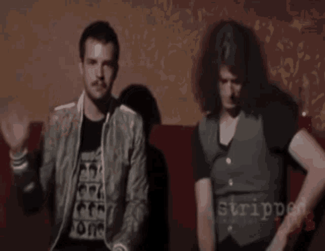 two men are sitting on a couch and one of them is wearing a vest that says strippers