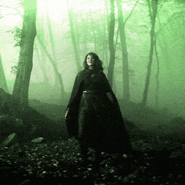a woman in a black cape is walking through a forest