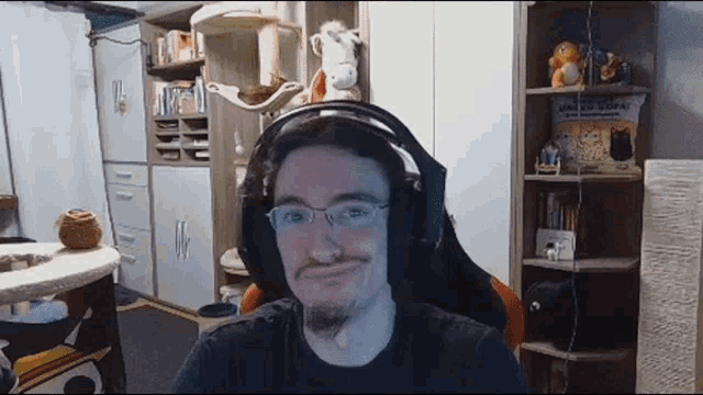 a man wearing headphones and glasses is sitting in a room with a cat tree in the background .