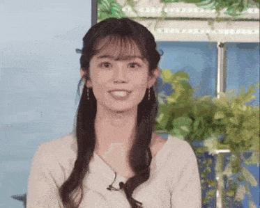 a woman with long hair and bangs is smiling and looking at the camera in front of a plant .