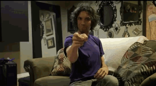 a man in a purple shirt is sitting on a couch pointing at the camera