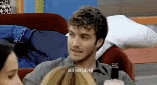 a man with curly hair is sitting on a couch with two women and a bottle of beer .