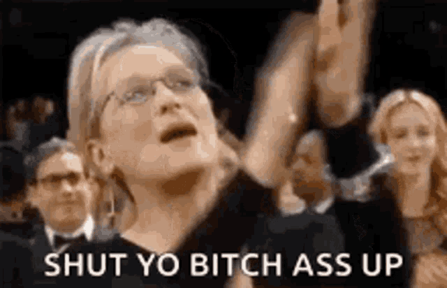 a woman wearing glasses is standing in front of a crowd and yelling at them .