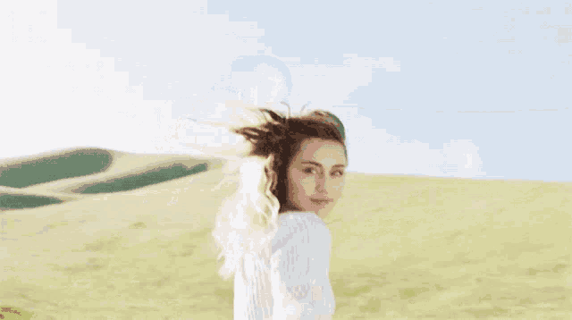 a woman in a white sweater stands in a field with her hair blowing in the wind