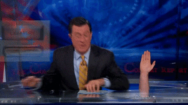 a man in a suit and tie is sitting at a desk with his hand up in front of a screen that says cnn