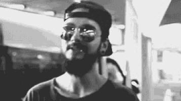 a man with a beard wearing sunglasses and a baseball cap is smoking a cigarette .