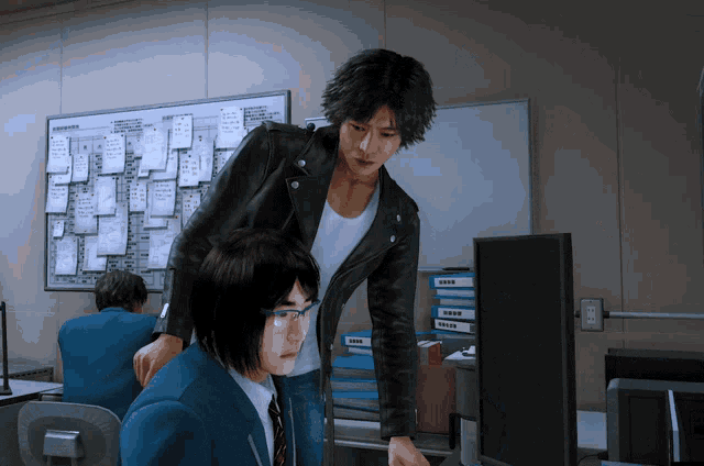 a man in a leather jacket stands next to a man in a suit looking at a computer monitor