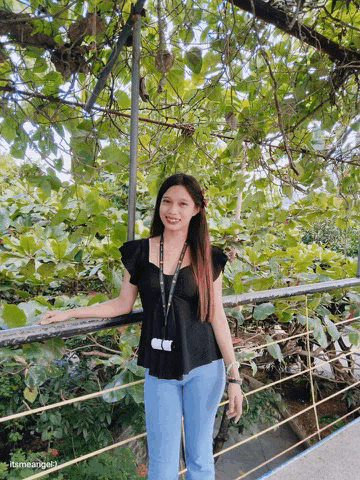 a woman in a black top and blue jeans stands in front of a lush green forest