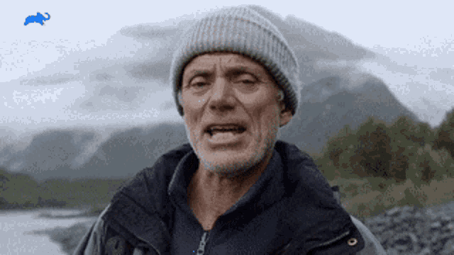 a man wearing a beanie and a jacket is standing in front of mountains