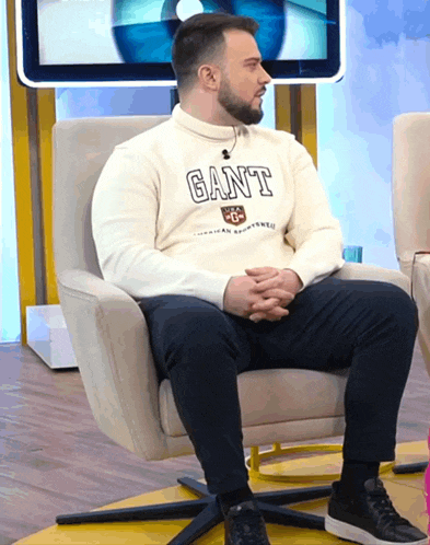 a man wearing a white sweater with the word gant on it sits in a chair