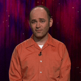 a man in an orange shirt with a microphone on his neck stands in front of a red curtain