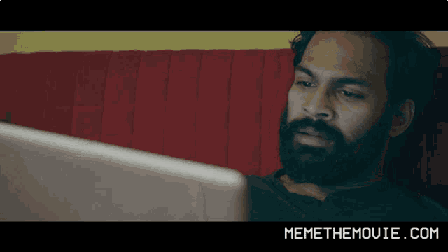 a man laying on a red couch looking at a laptop screen with memethemovie.com written below him