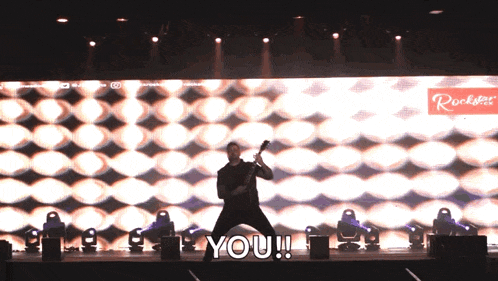 a man playing a guitar on a stage with the words " you " behind him