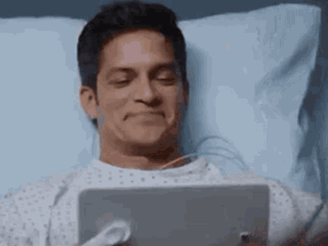 a man is laying in a hospital bed looking at a tablet computer .
