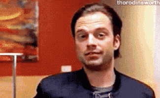 a man in a black jacket is standing in front of a red wall and making a funny face .