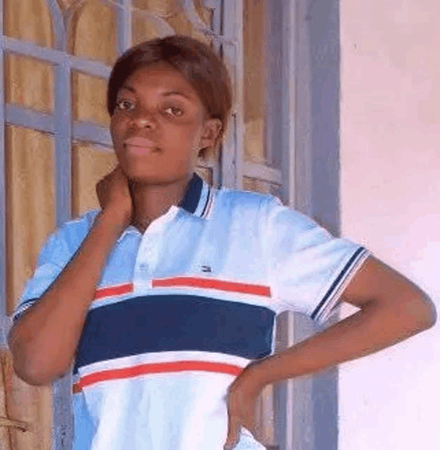 a young woman in a blue and white striped polo shirt is standing in front of a window with her hand on her chin .