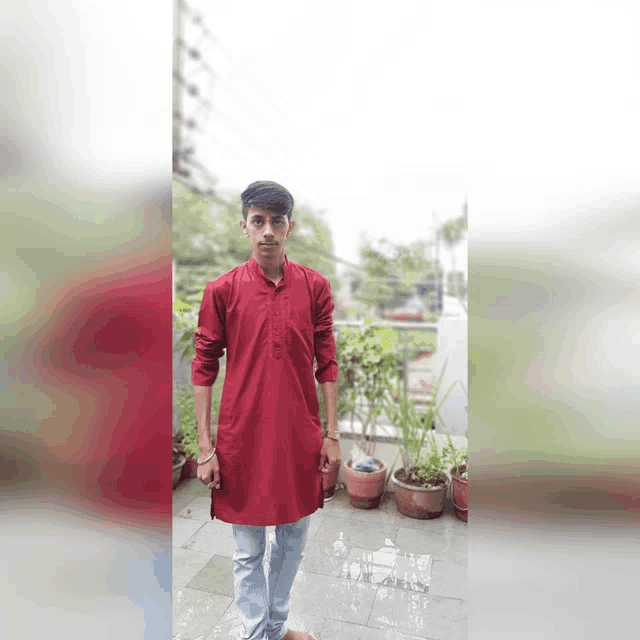 a young man wearing a red shirt and blue jeans stands in front of potted plants