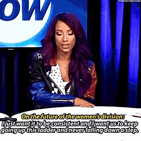 a woman with purple hair is sitting at a table in front of a television talking about the future of the women 's division .