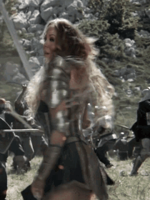a woman in armor stands in a field with a sword in her hand