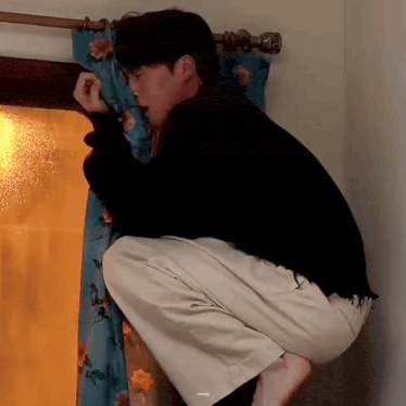 a man in a black sweater and white pants is squatting down in front of a window .