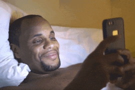 a shirtless man is laying in bed looking at a cell phone
