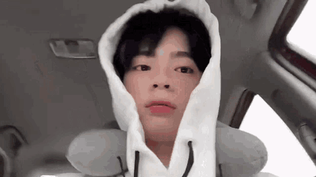 a young man wearing a white hoodie is sitting in a car with a pillow on his neck .