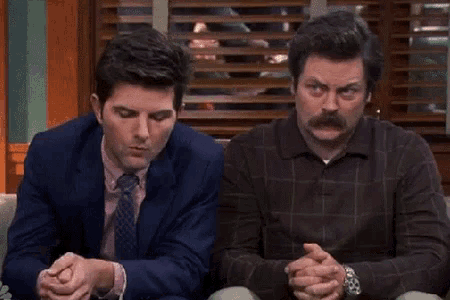 two men are sitting next to each other with their hands folded and looking at each other .