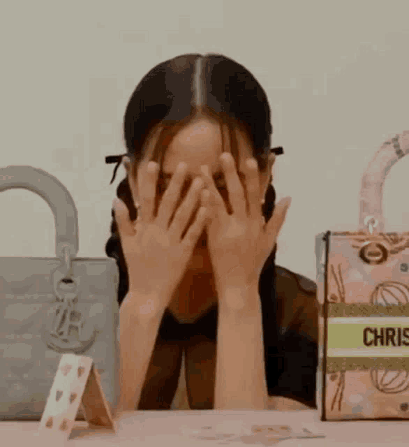 a woman is covering her face with her hands while sitting at a table with handbags .