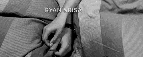 ryan and risa are holding hands while laying on a bed .