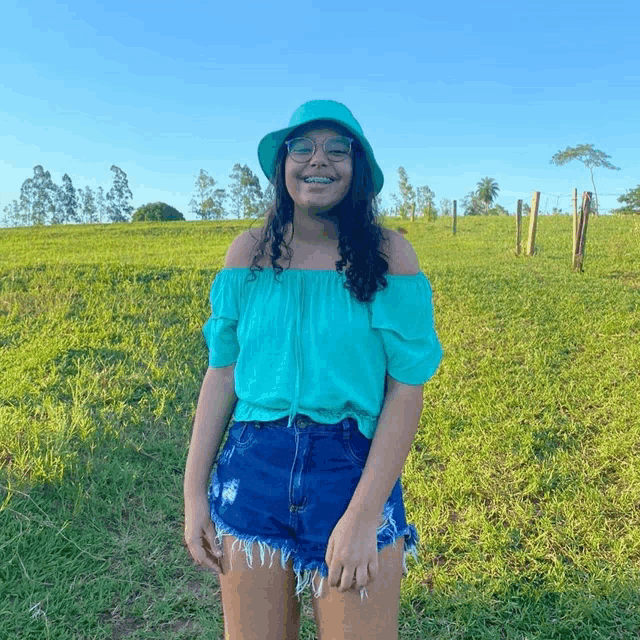 a girl wearing a green off the shoulder top and blue shorts is standing in a grassy field