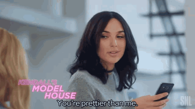 a woman is talking to another woman while holding a cell phone and saying you 're prettier than me .