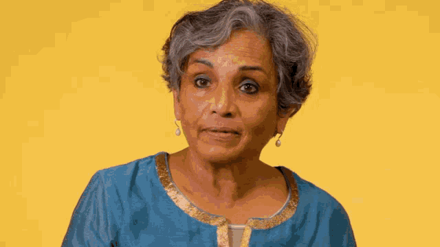a woman wearing a blue shirt and earrings looks at the camera with a yellow background