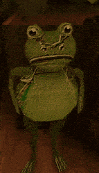 a green frog with a sad face is sitting on a wooden floor