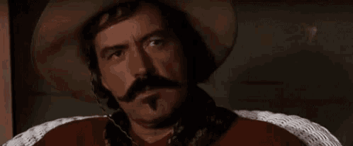 a man with a hat and mustache is sitting in a wicker chair and saying `` well '' .
