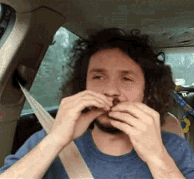 a man with curly hair and a beard is sitting in a car eating a hamburger