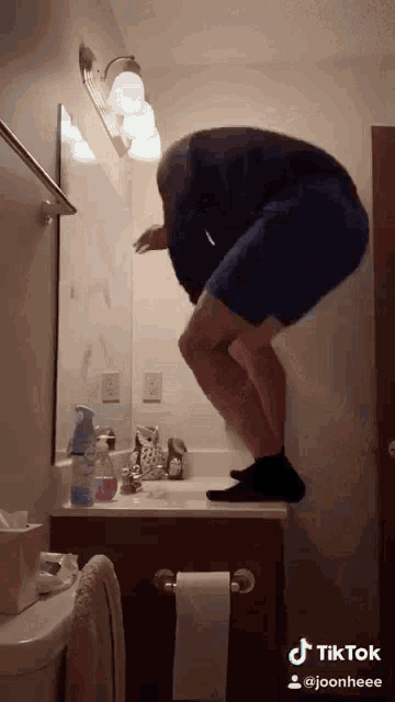 a man is squatting on a bathroom sink while looking at himself in the mirror .