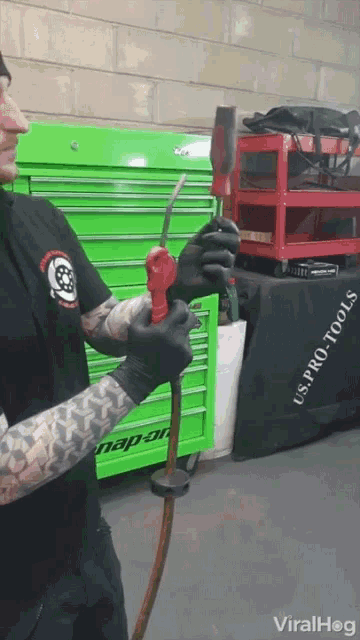 a man wearing black gloves is holding a tool in front of a green snap-on toolbox