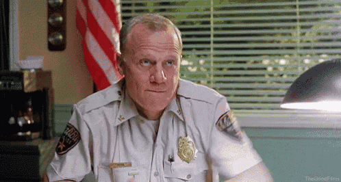 a man in a police uniform is sitting at a desk in front of a window with blinds .