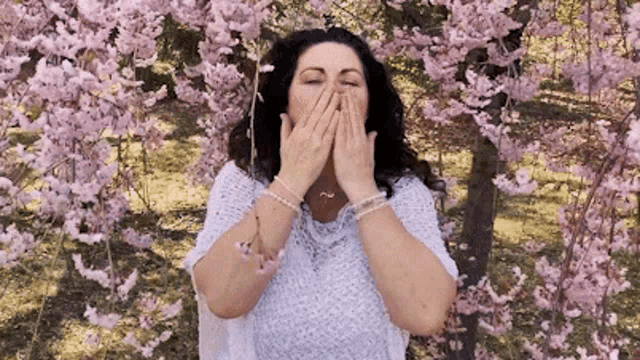 a woman covering her mouth with her hands while standing in front of a tree with pink flowers