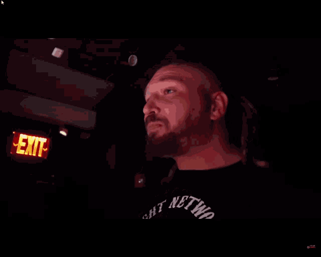 a man in a black shirt with the word network on it stands in front of a red exit sign